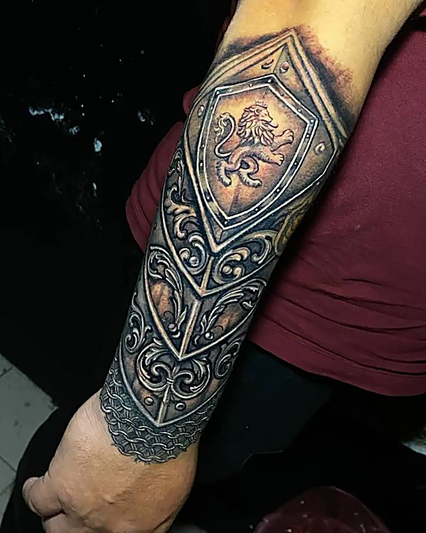 Antebrazo con un brazalete de armadura tatuado: escudo con león, filigrana y reflejos metálicos, en negro y gris